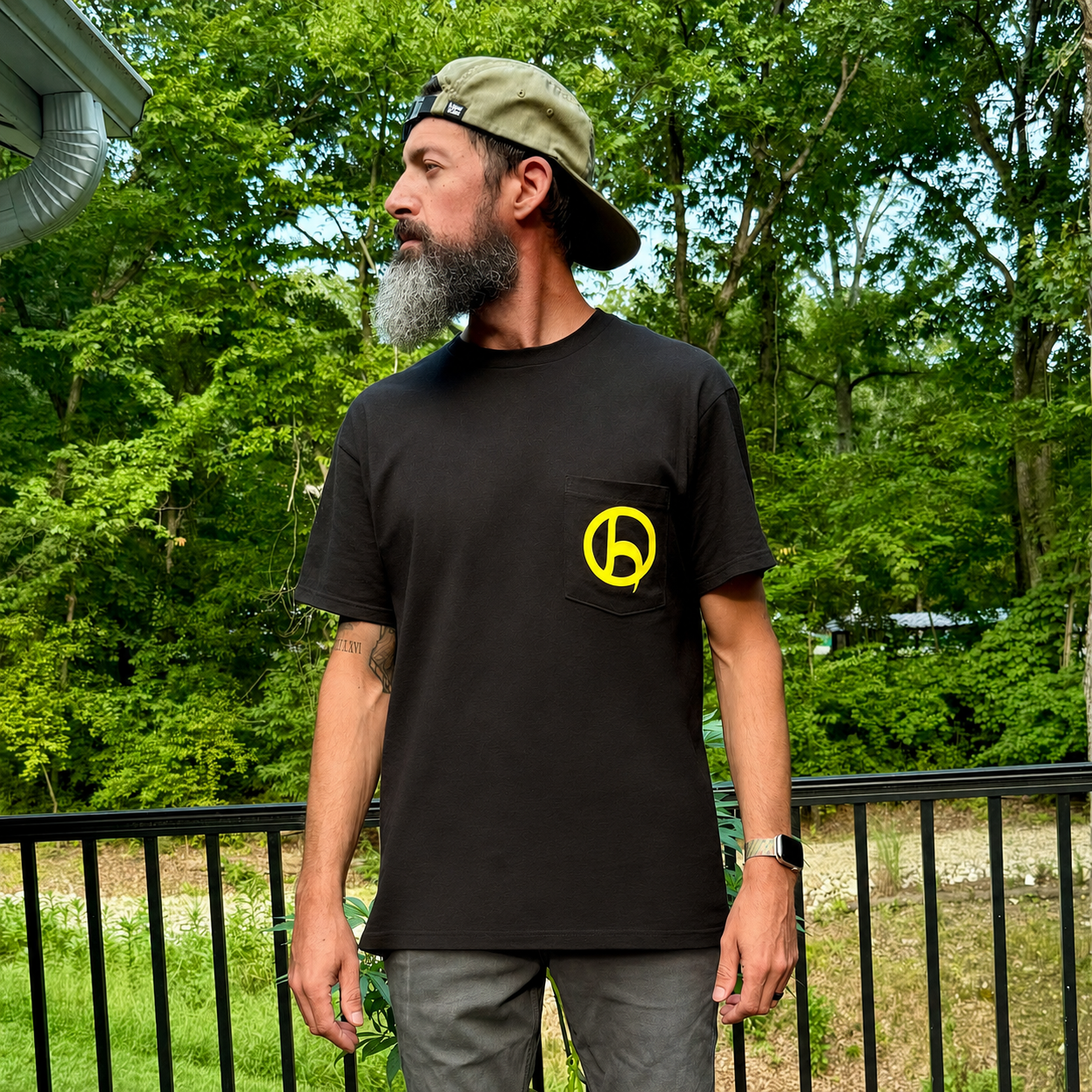 A man with a gray beard wearing a black crewneck HempHooks logo pocket t-shirt with a yellow circular logo on the chest, paired with a khaki cap and gray pants, standing outdoors on a balcony with a green forest background.