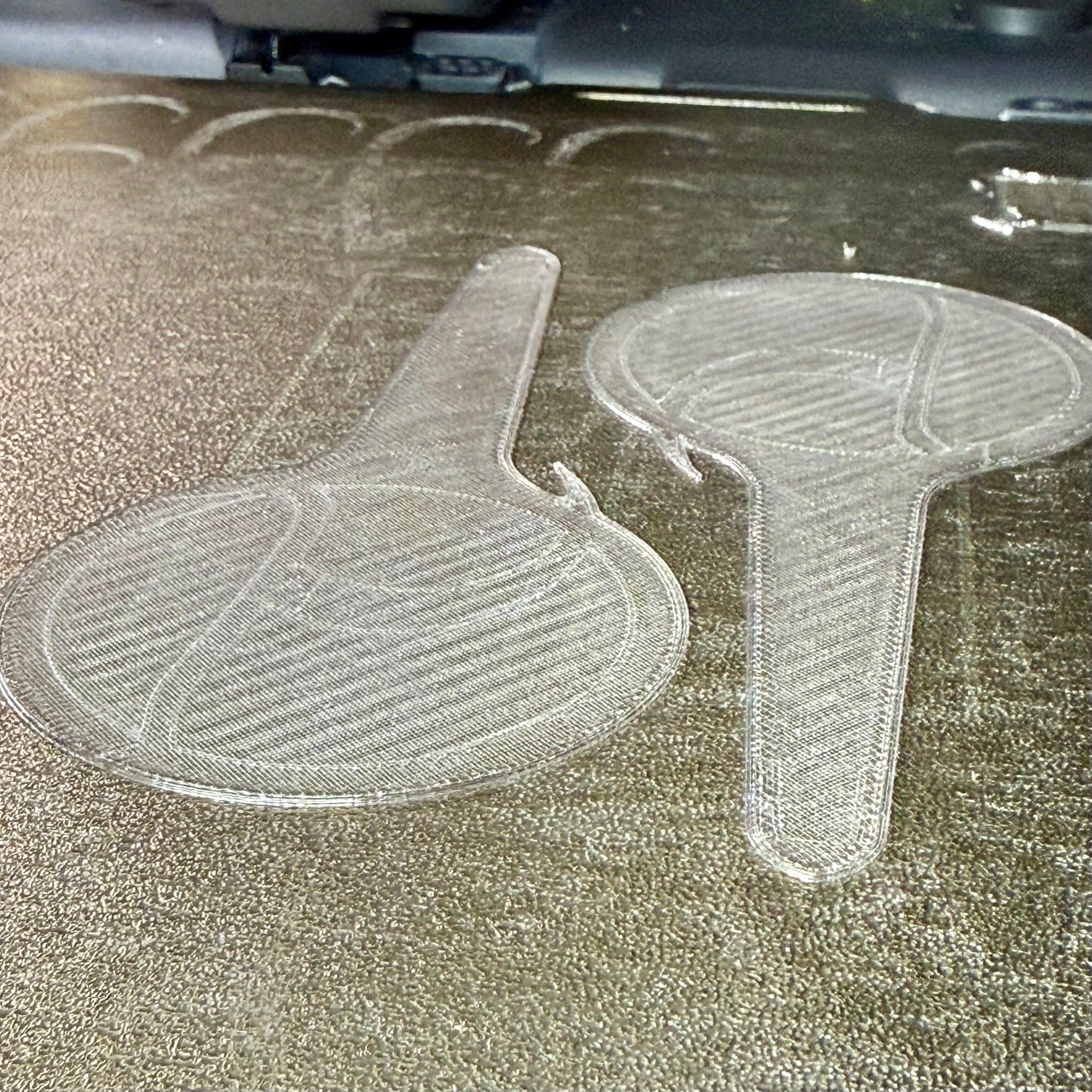 Two clear, 3D-printed paddle-shaped tags or labels resting on a textured, metallic surface.